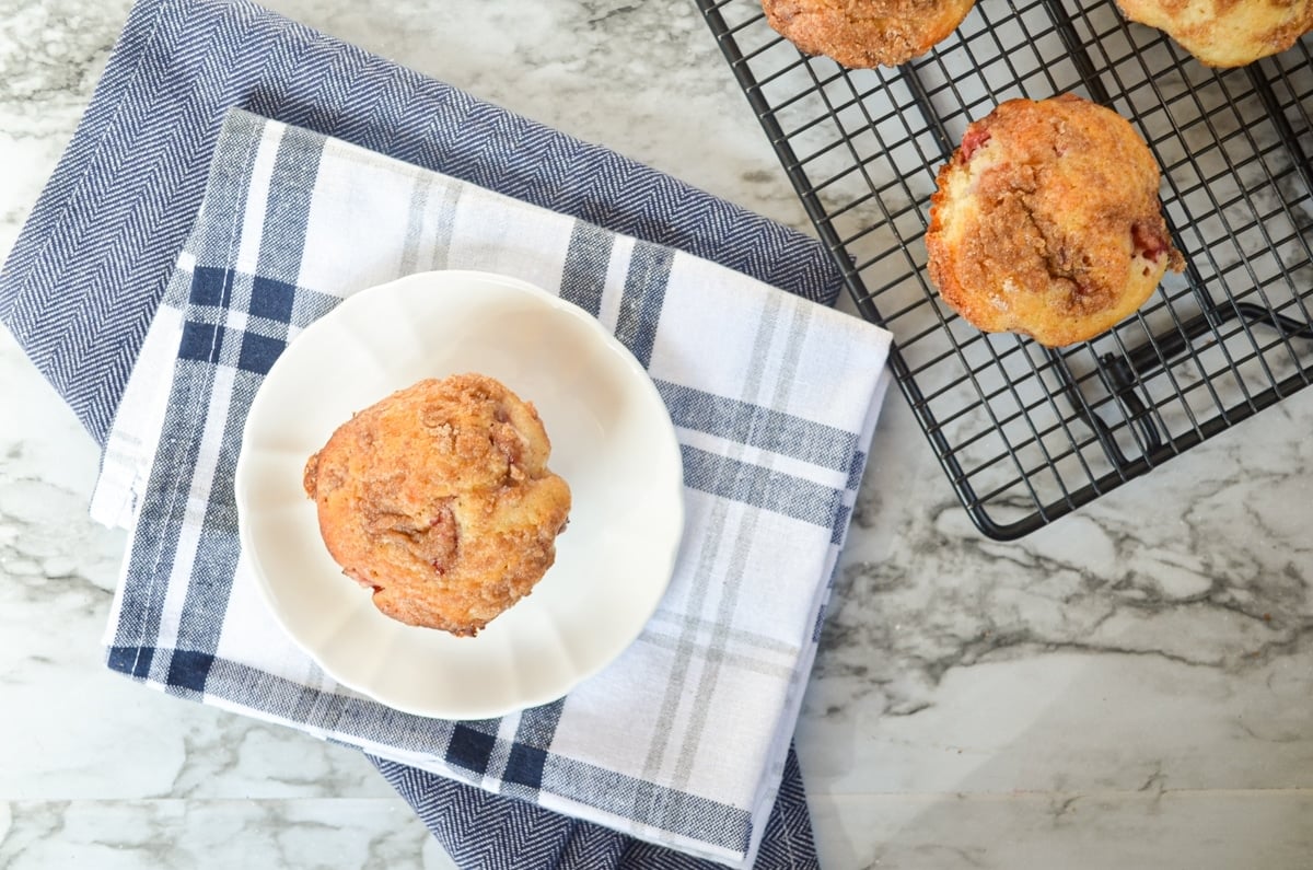 An overhead shot of a muffin on a plate, resting on cloth napkins. A cooling rack to the right with 3 muffins in view.