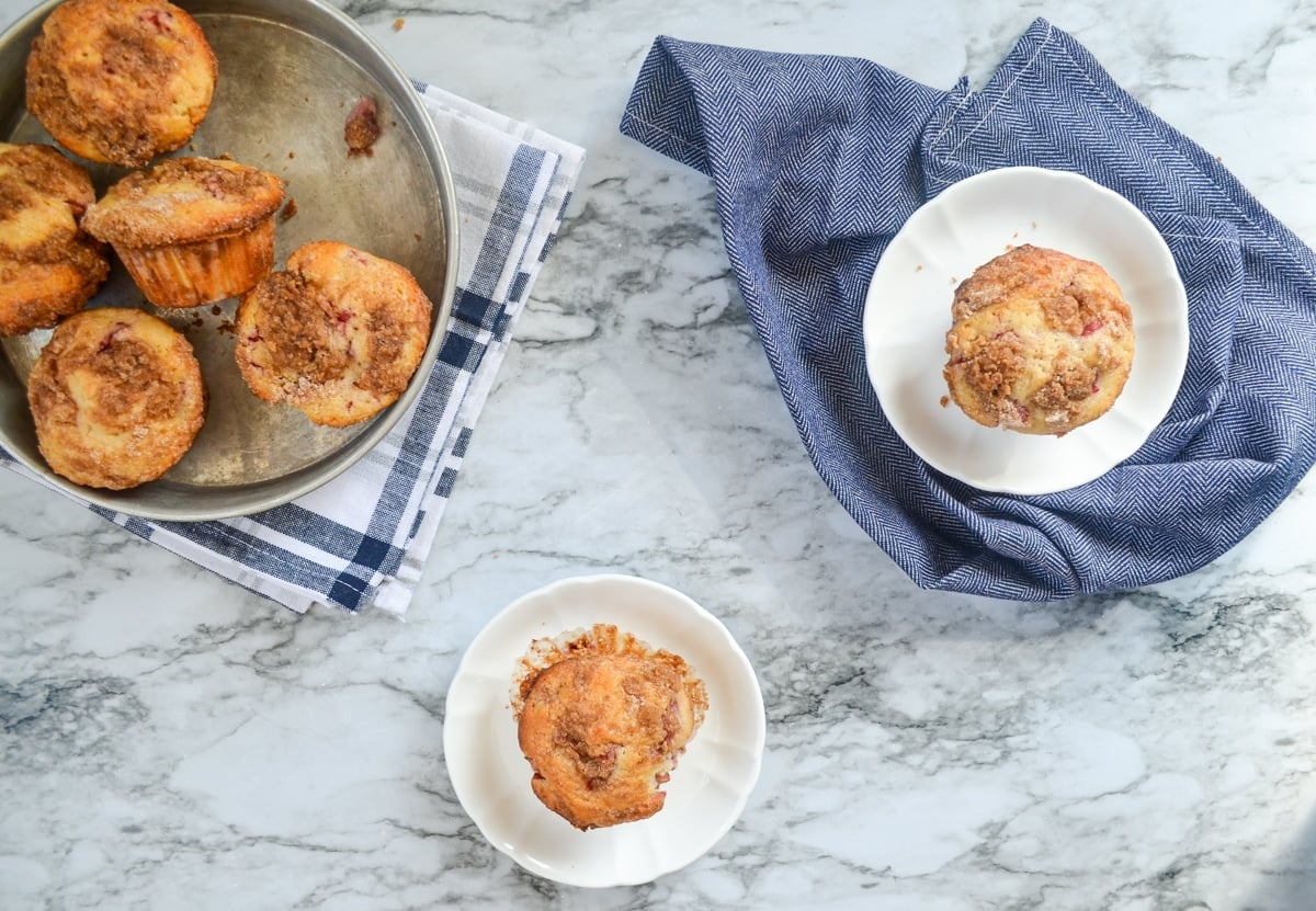 An overhead view of strawberry buttermilk muffins. One is on a white plate on the counter, another on a white plate on a napkin, and a few more in a pan to the left hand side.