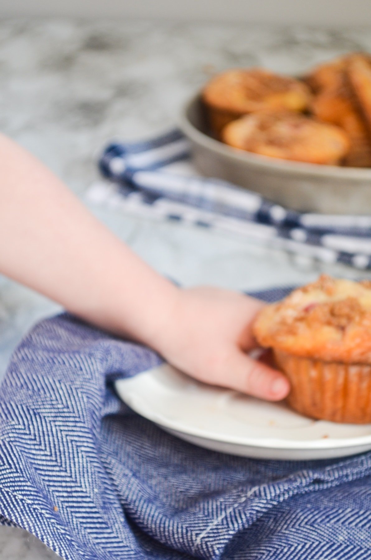 A muffin on a white plate, resting on a blue napkin and being grabbed by a toddler.