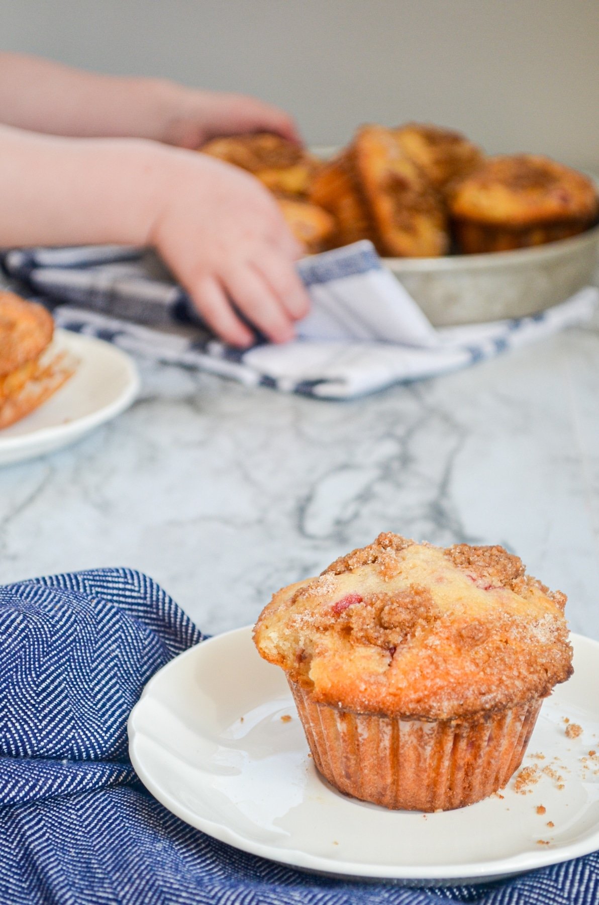 A strawberry buttermilk muffin on a white plate, resting on a blue napkin. In the background, a child lifts up a tray of muffins.