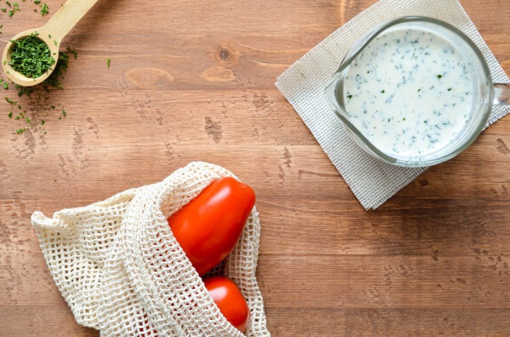 An overheat shot of buttermilk ranch dressing, a few tomatoes in a reusable produce bag, and a measuring spoon of dried parsley.