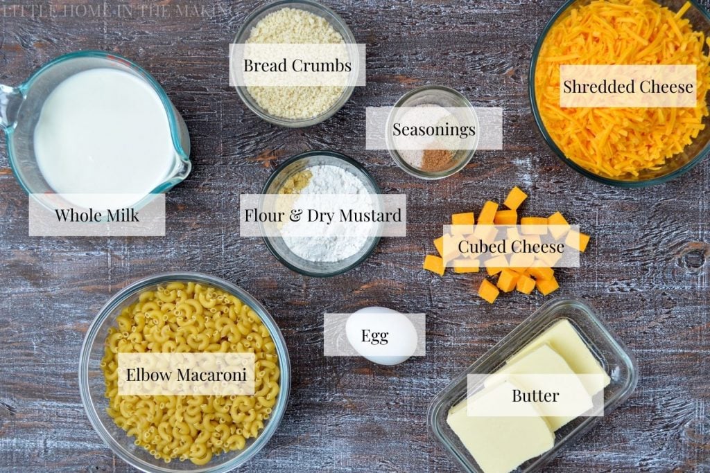 All of the ingredients needed to make macaroni from scratch: cheese, seasonings, flour, mustard powder, breadcrumbs, butter, egg, macaroni, and milk.