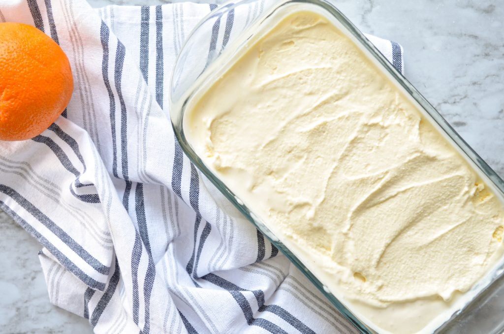 A glass loaf pan with orange creamsicle ice cream inside, resting on a blue and white tea towel.