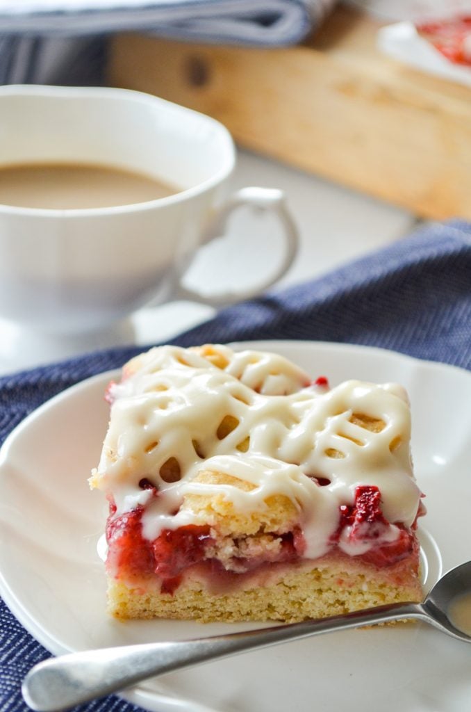 A strawberry crumb bar on a white plate, with a small cup of coffee in the background.