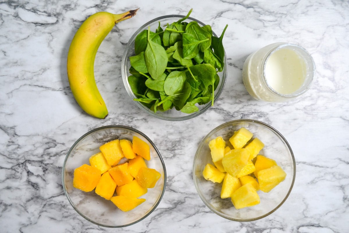 A bowl of spinach, another of mango, another of pineapple, and with a banana on the side.
