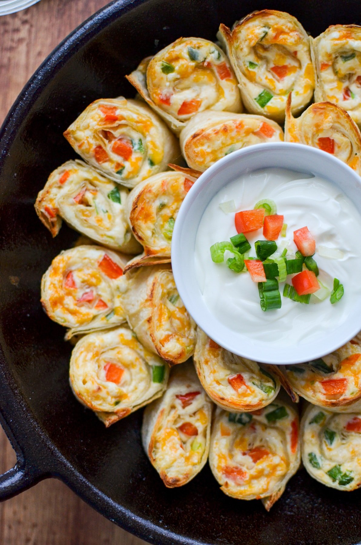 A top down shot of a cast iron skillet with jalapeno and cream cheese pinwheels in them. A bowl of sour cream in the center.