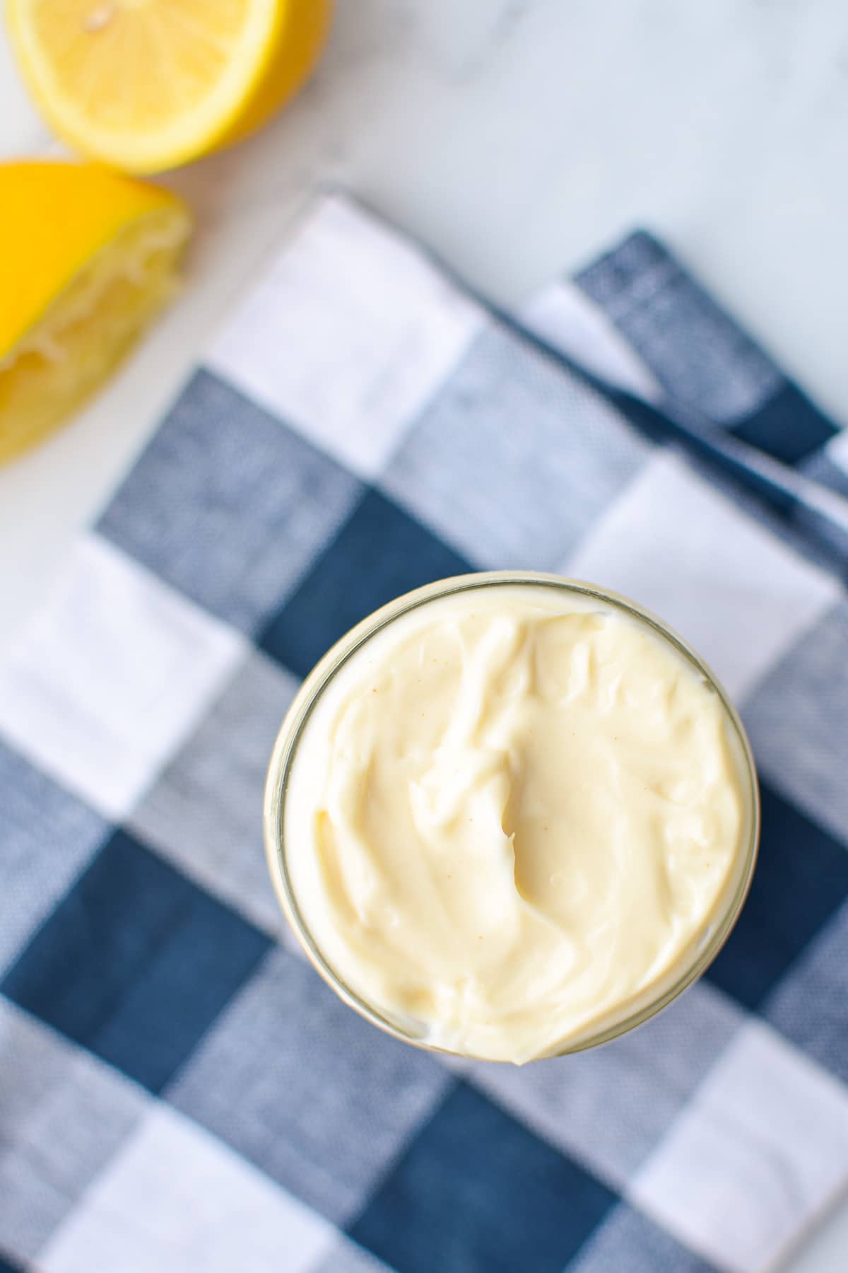 An overhead shot of a jar of homemade avocado oil mayo. A blue check napkin and fresh squeezed lemon in the background.