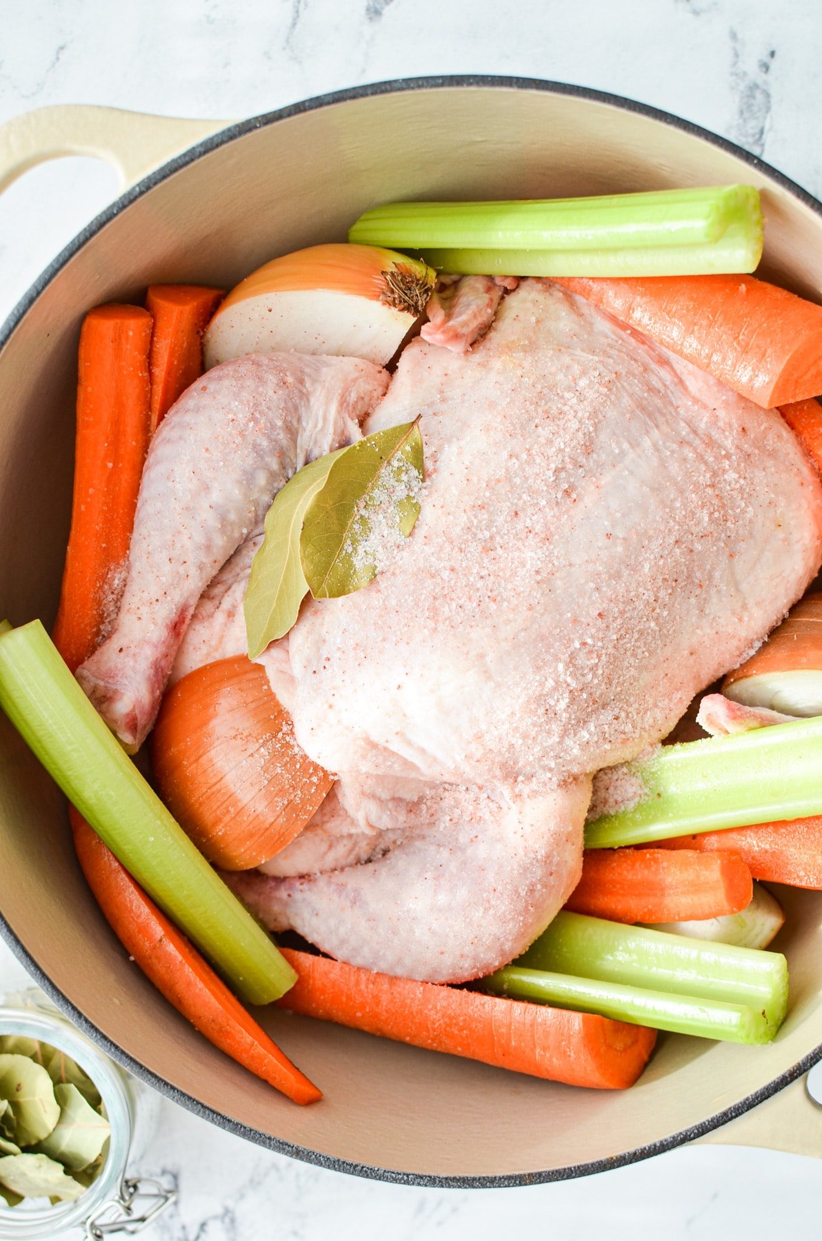 A whole chicken inside of a dutch oven, surrounded by vegetables and seasoned with salt.