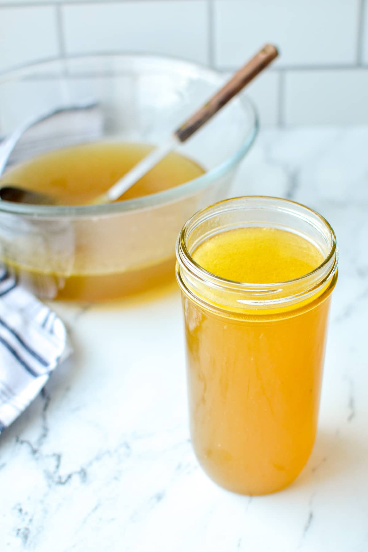 A glass jar full of homemade chicken broth, with a glass bowl and ladle in the background.