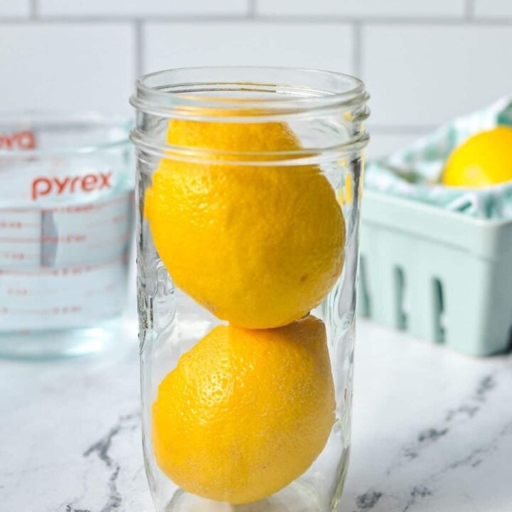A jar filled with lemons, with water and a container of lemons in the background.