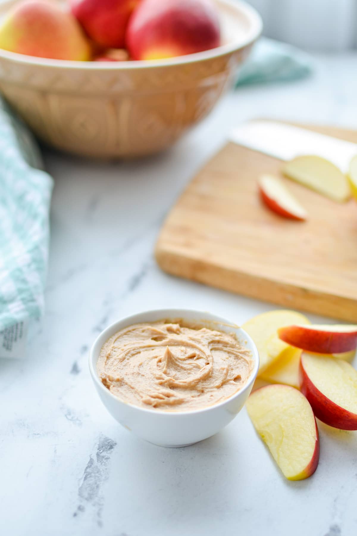 A small bowl of peanut butter yogurt dip, with sliced apples in the foreground and a bowl in the background.