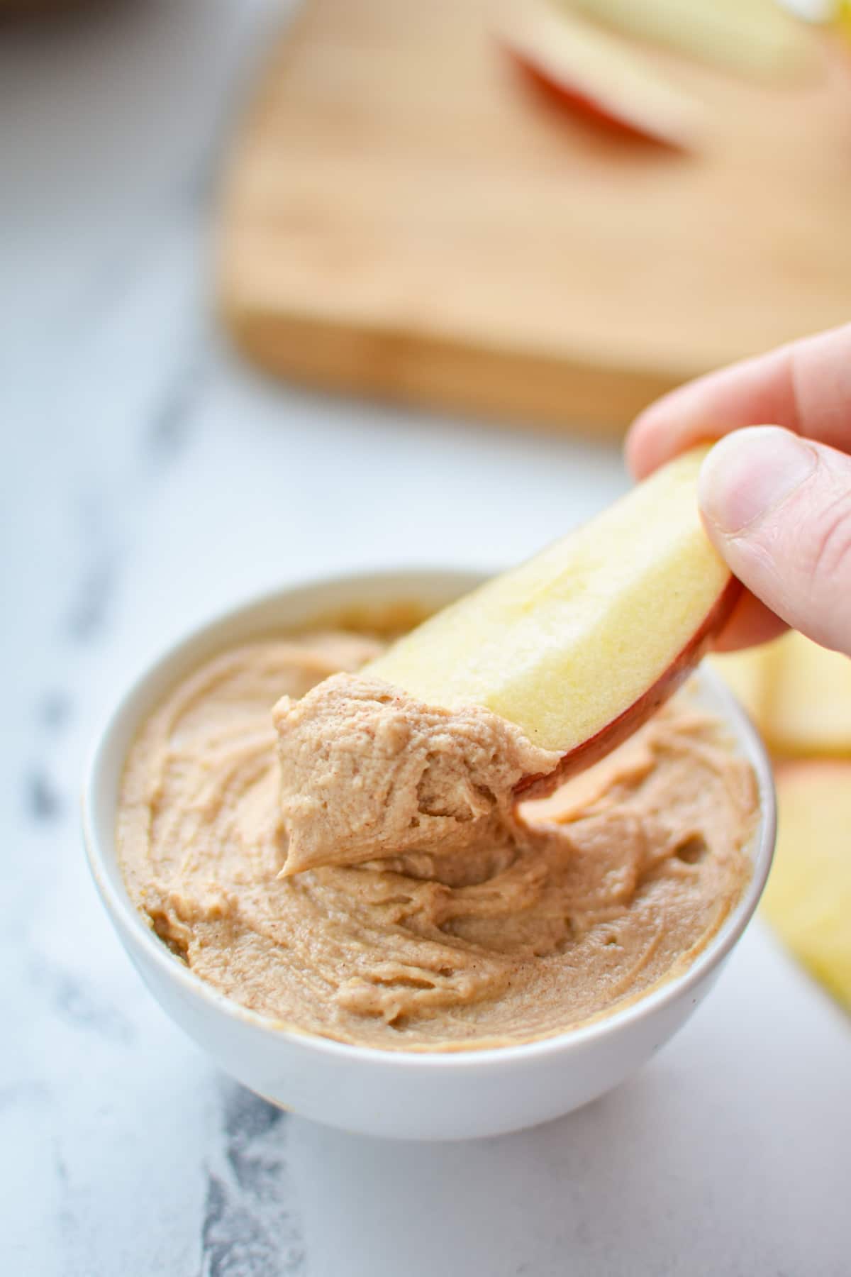 Dipping an apple slice into a peanut butter yogurt dip, with a cutting board in the background.