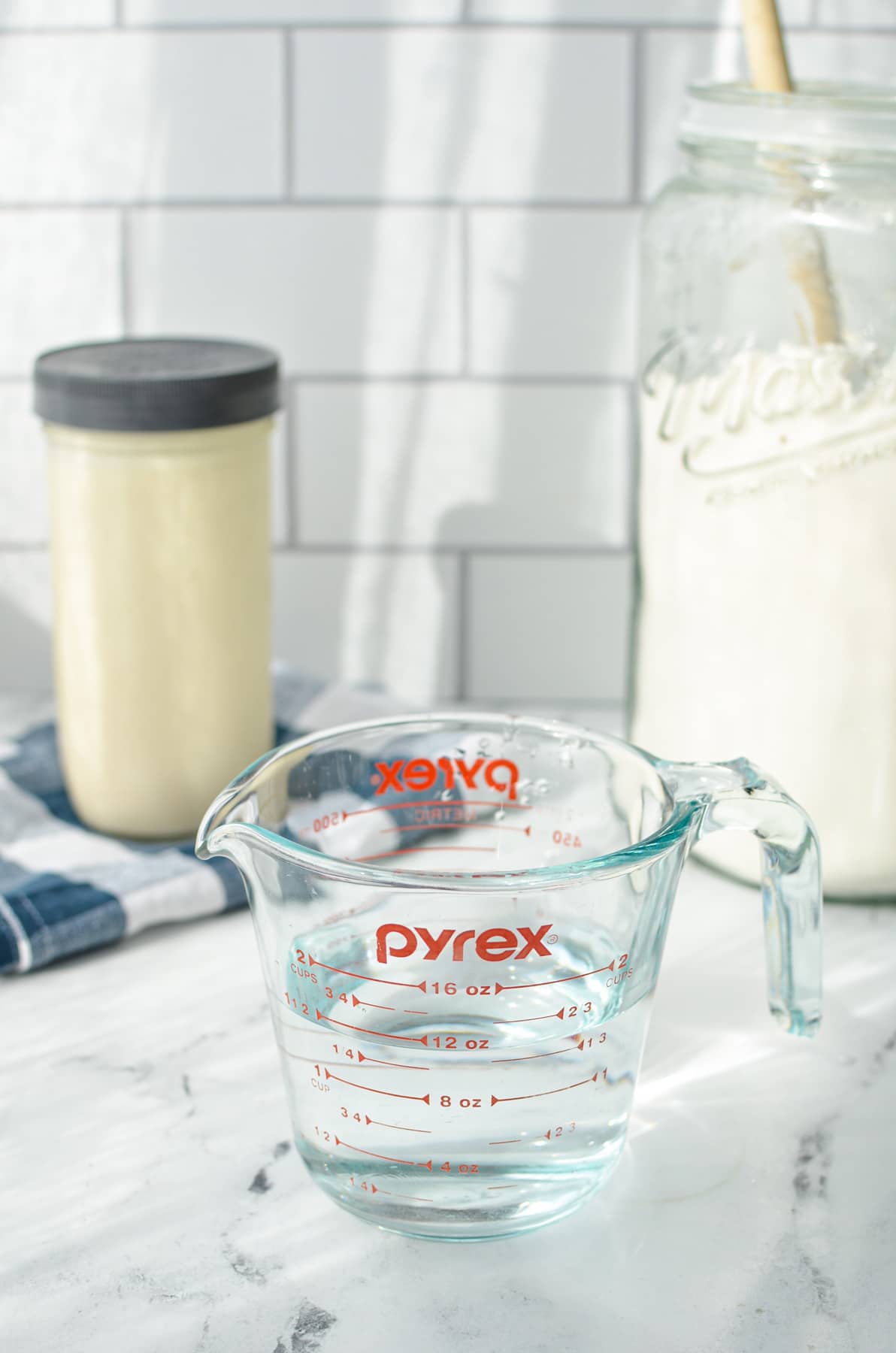 A counter with a glass measuring cup of water, canister of flour, and a jar of sourdough starter.