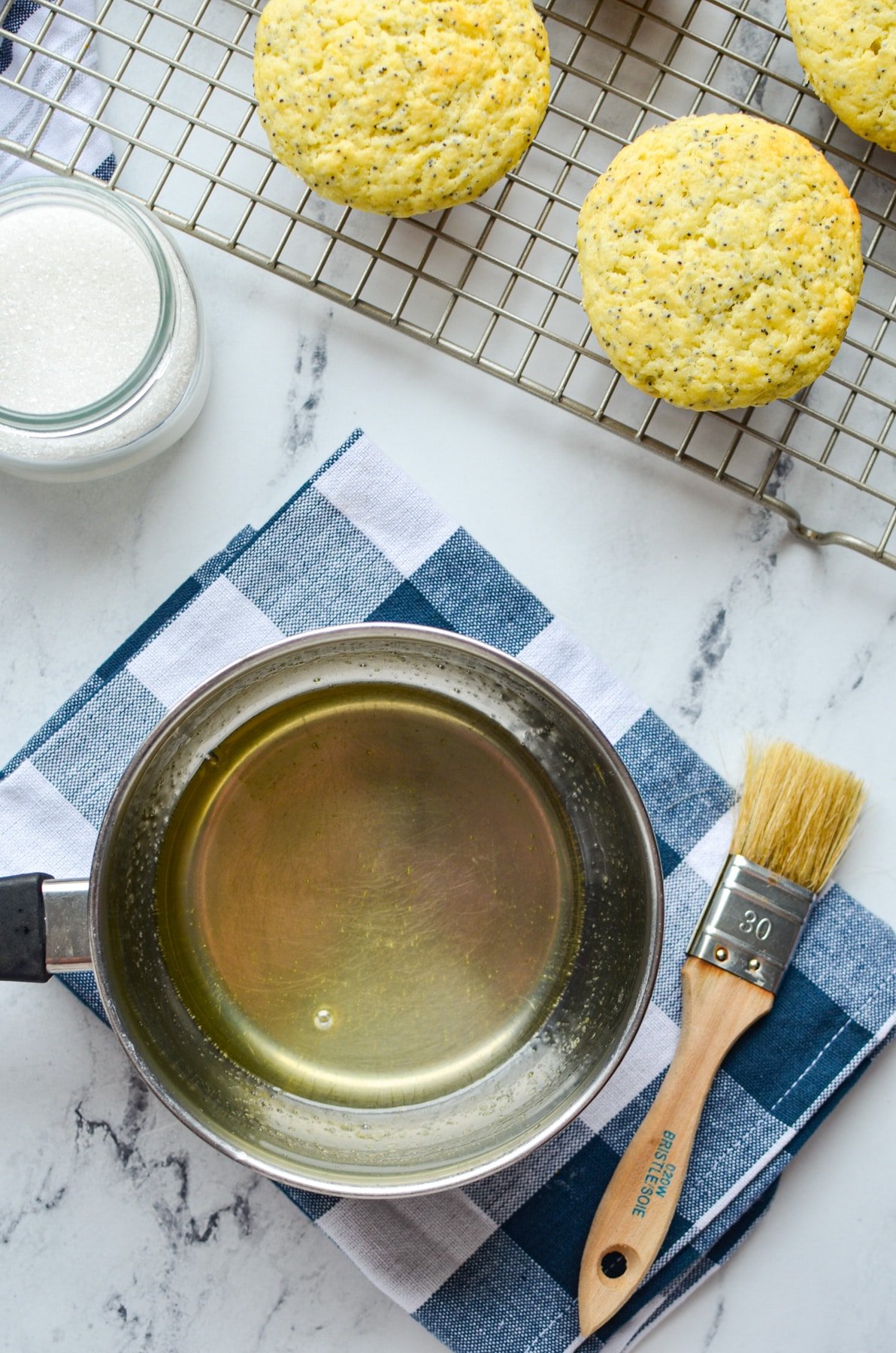 A small saucepan filled with a lemon glaze, with lemon poppy seed muffins in the background.