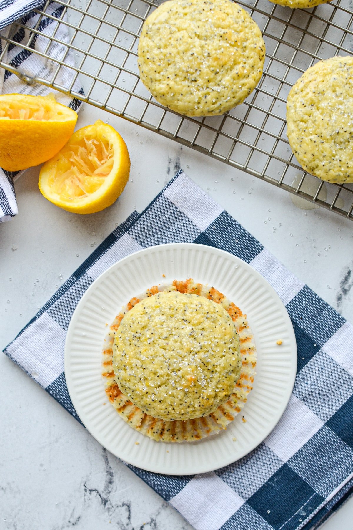 A lemon muffin on a blue check napkin, with a wire rack in the background and some squeezed lemons.