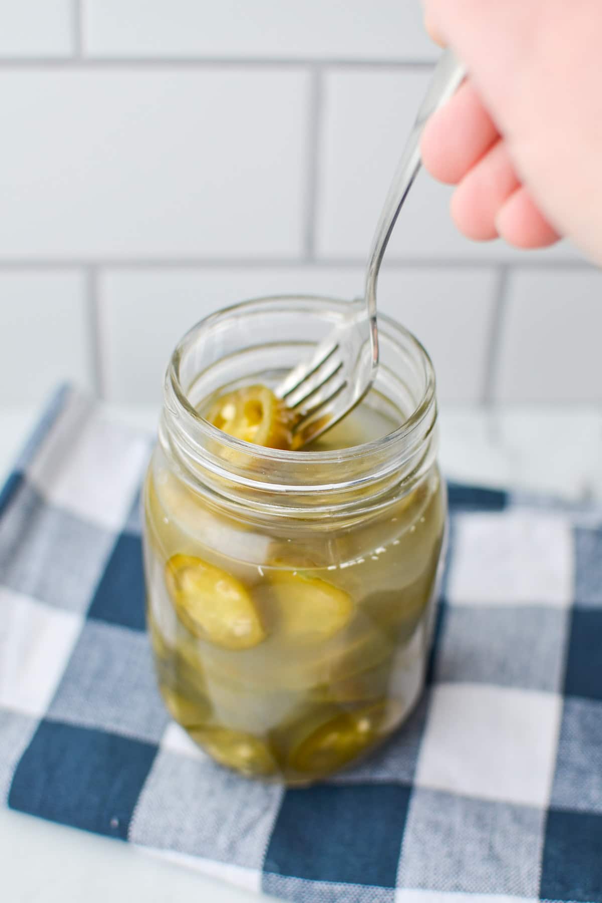A jar of fermented jalapenos on a blue check cloth; a hand using a fork to remove a ring.