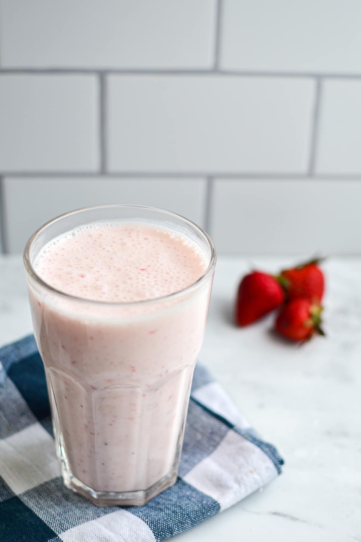 A glass of strawberry milk kefir on a blue check napkin, with strawberries in the background.