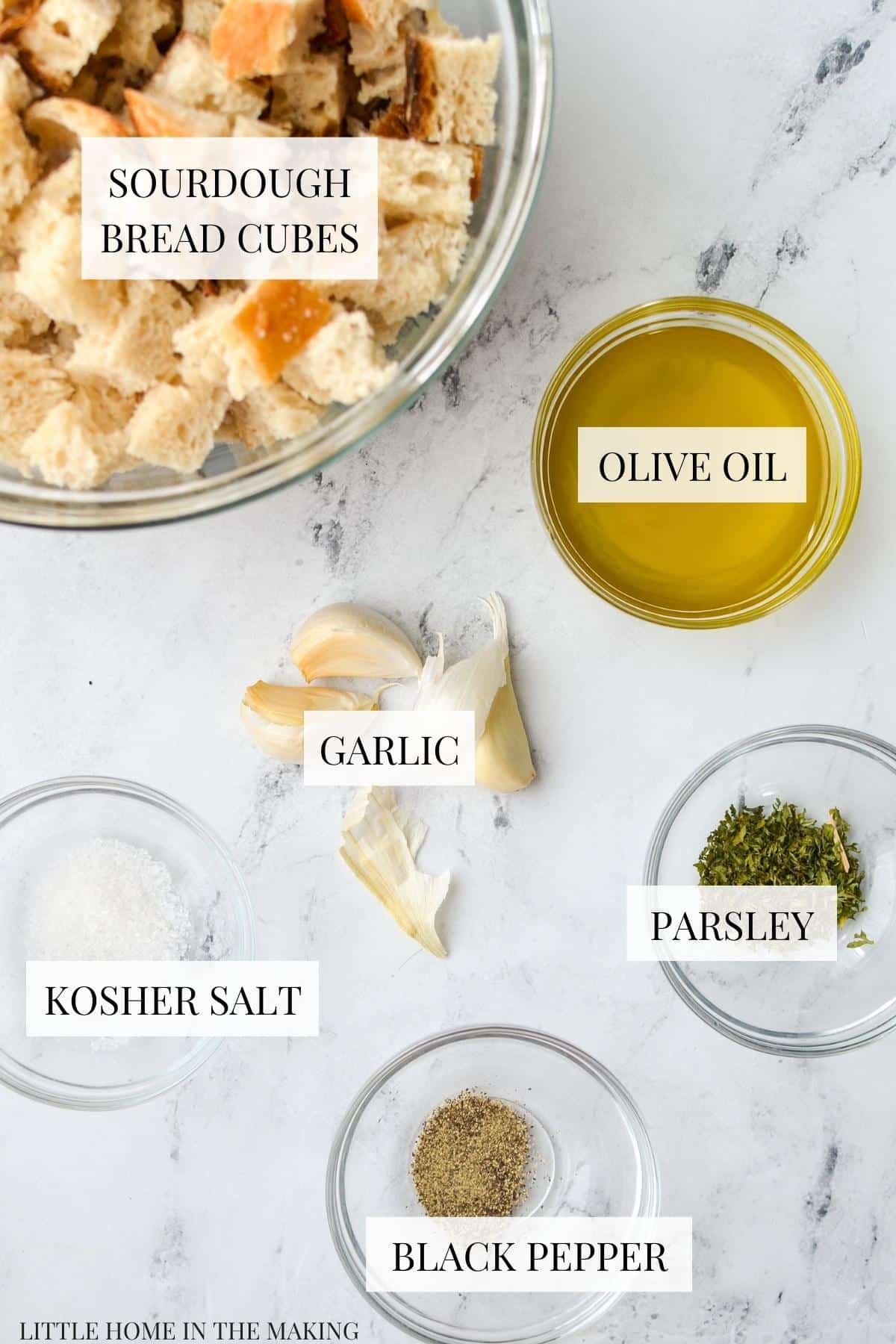 The ingredients needed to make sourdough croutons, including: sourdough bread cubes, olive oil, salt, pepper, parsley and garlic,