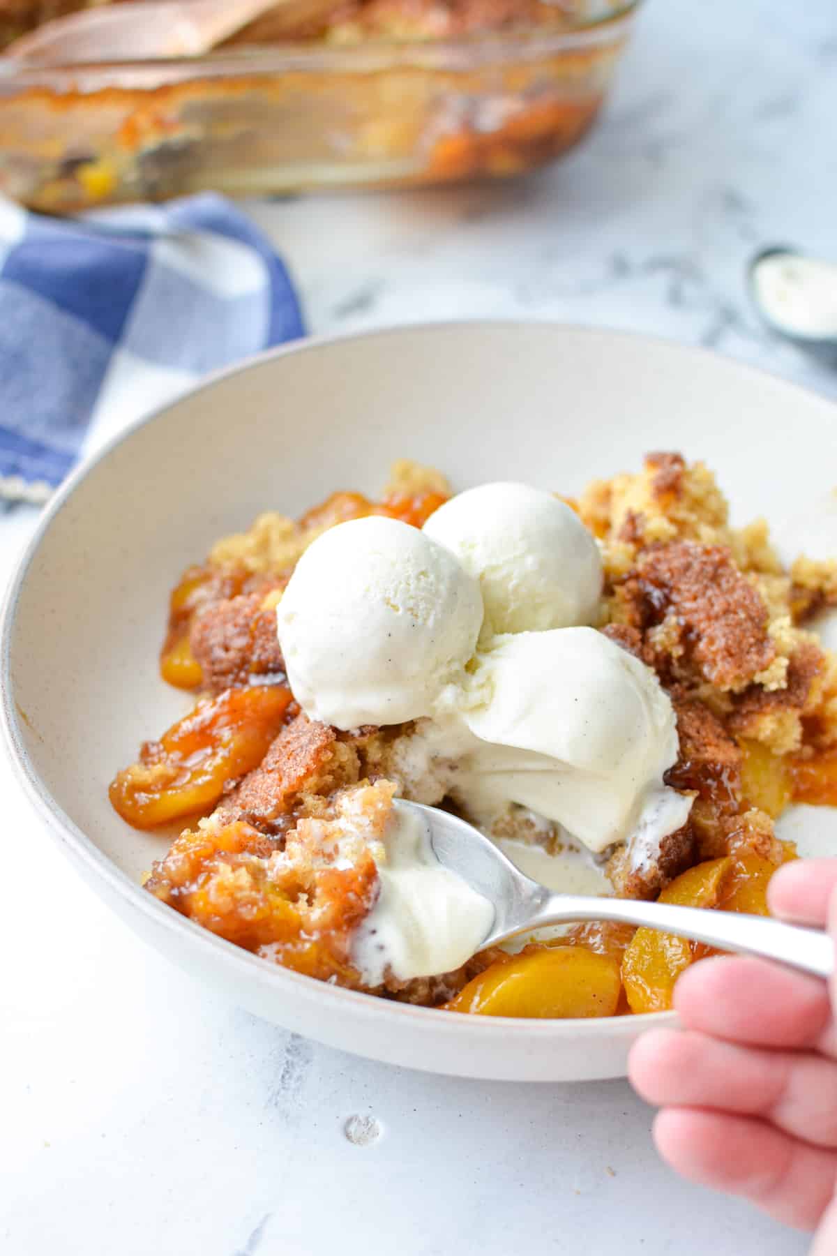 A spoon taking a portion of peach cobbler, served with several scoops of vanilla ice cream.