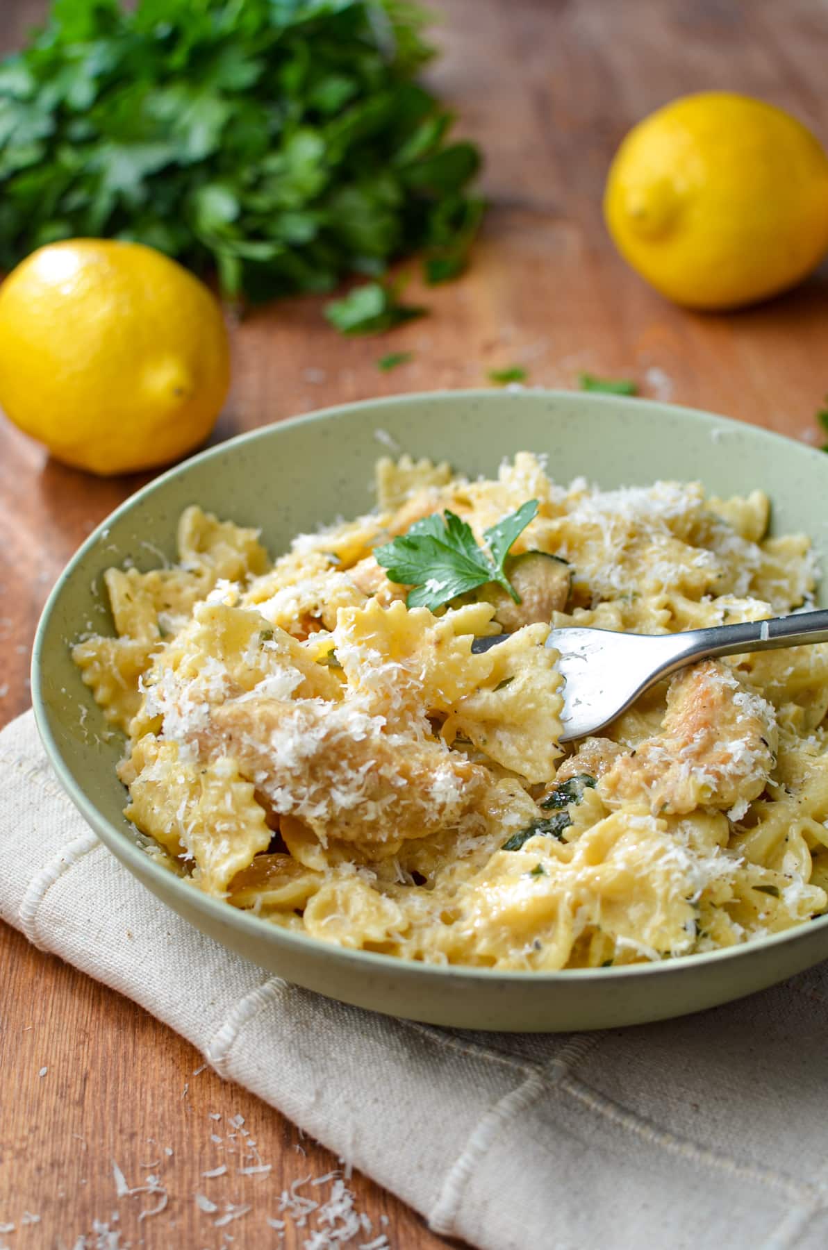 A bowl of lemon and chicken pasta with parsley and lemons in the background.