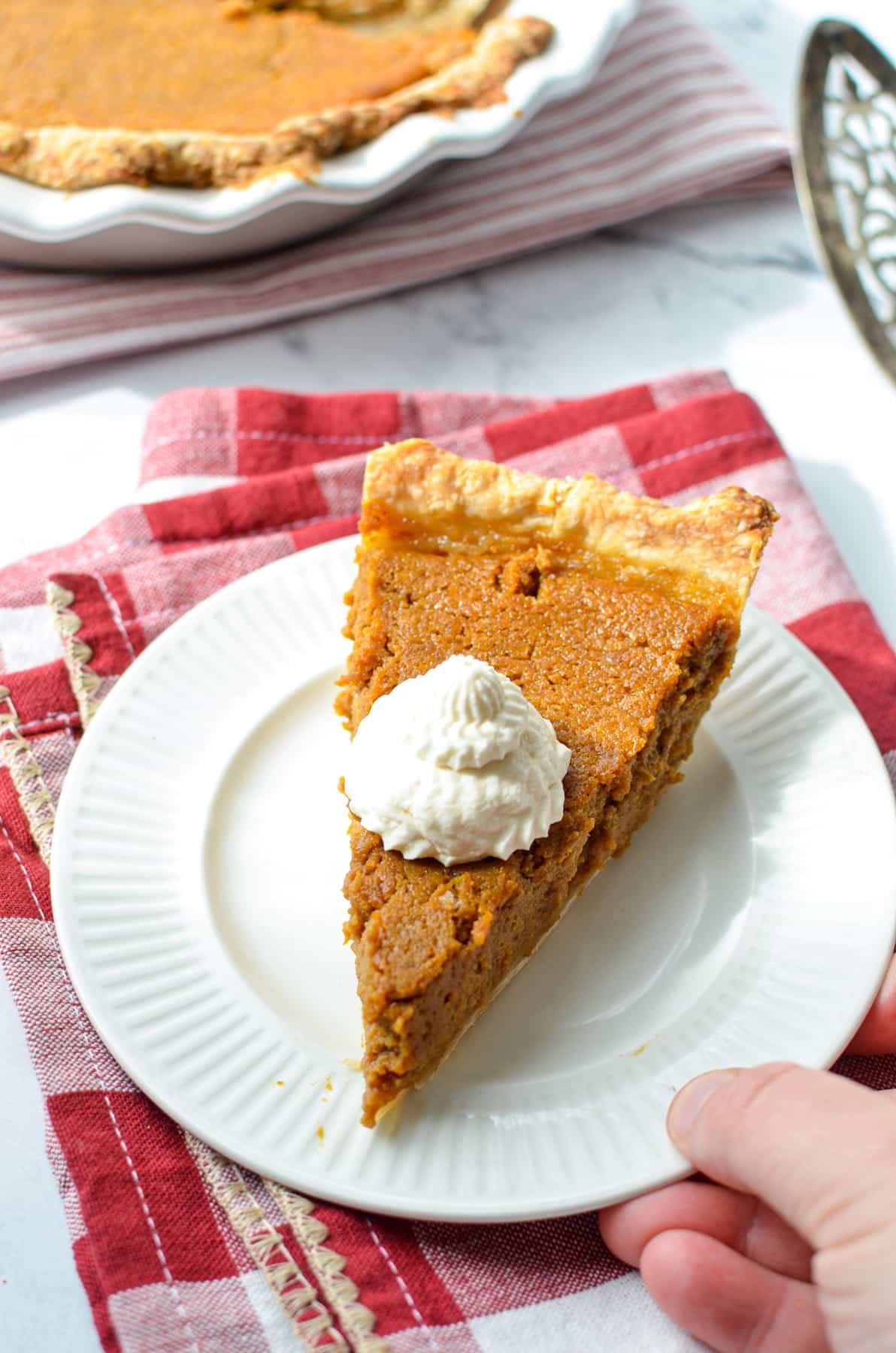 A slice of pumpkin pie with whipped cream, with the entire pie in the background.