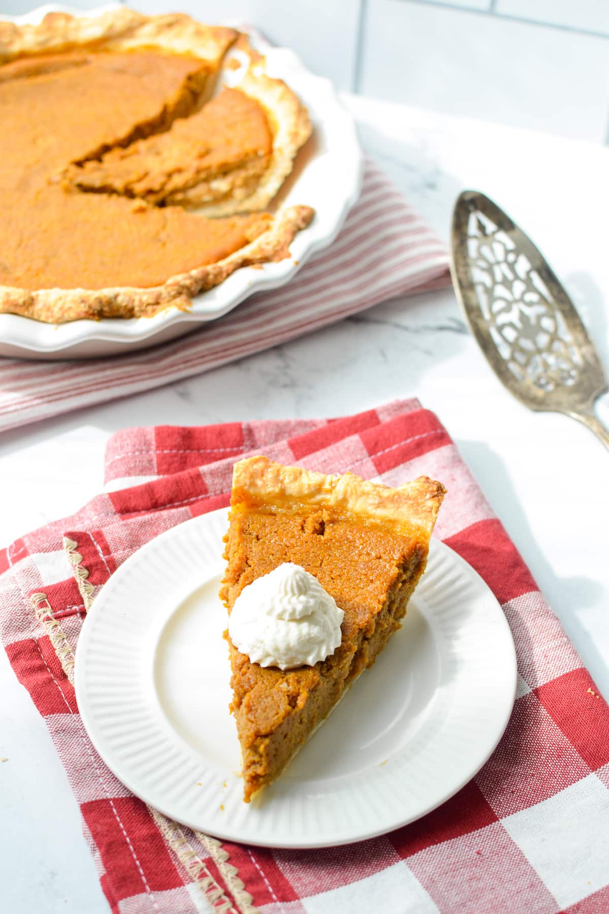 A slice of pumpkin pie with whipped cream, with a pie in the background.