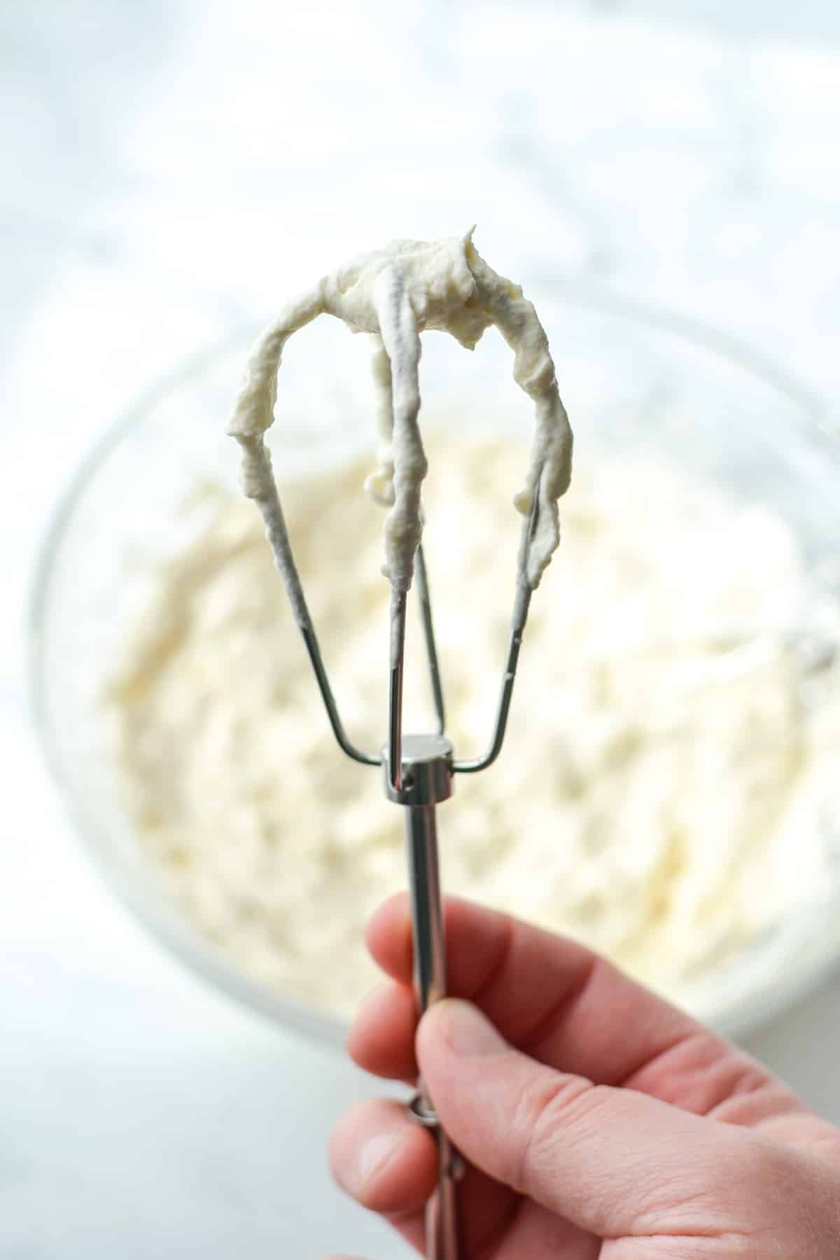 A hand holding a whisk covered with whipped cream.