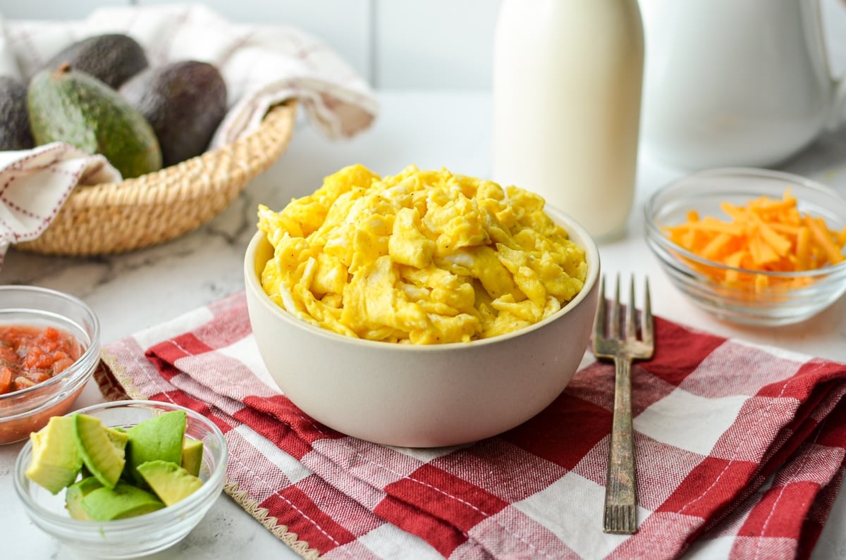 A bowl of scrambled eggs, with cheese, avocado, and salsa in prep bowls in the background.