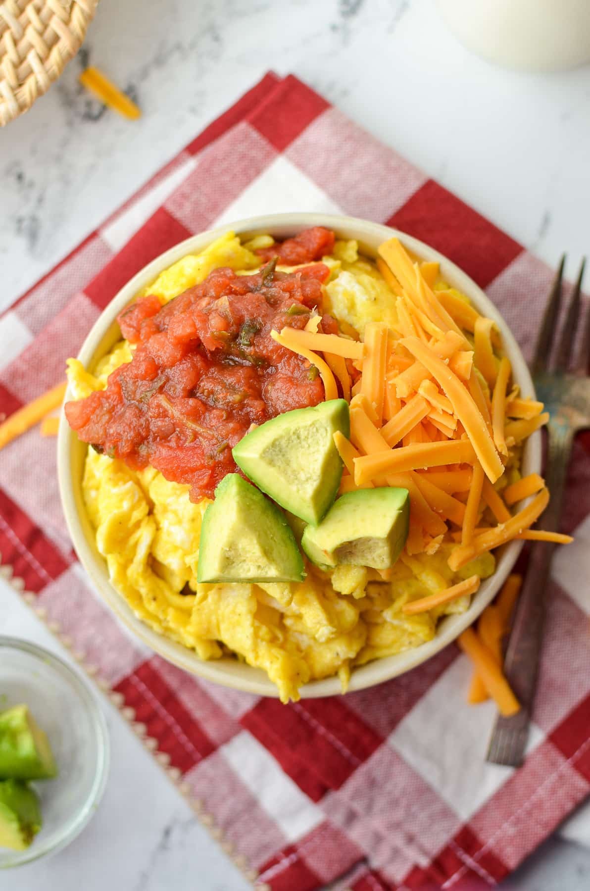 An overhead shot of a bowl with scrambled eggs, cheese, salsa, and avocado.