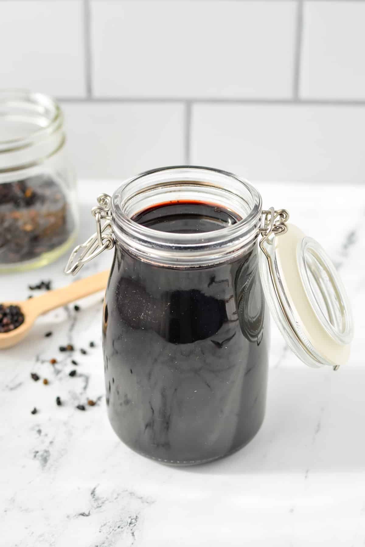 A jar filled with elderberry syrup, with elderberries scattered on the countertop behind it.