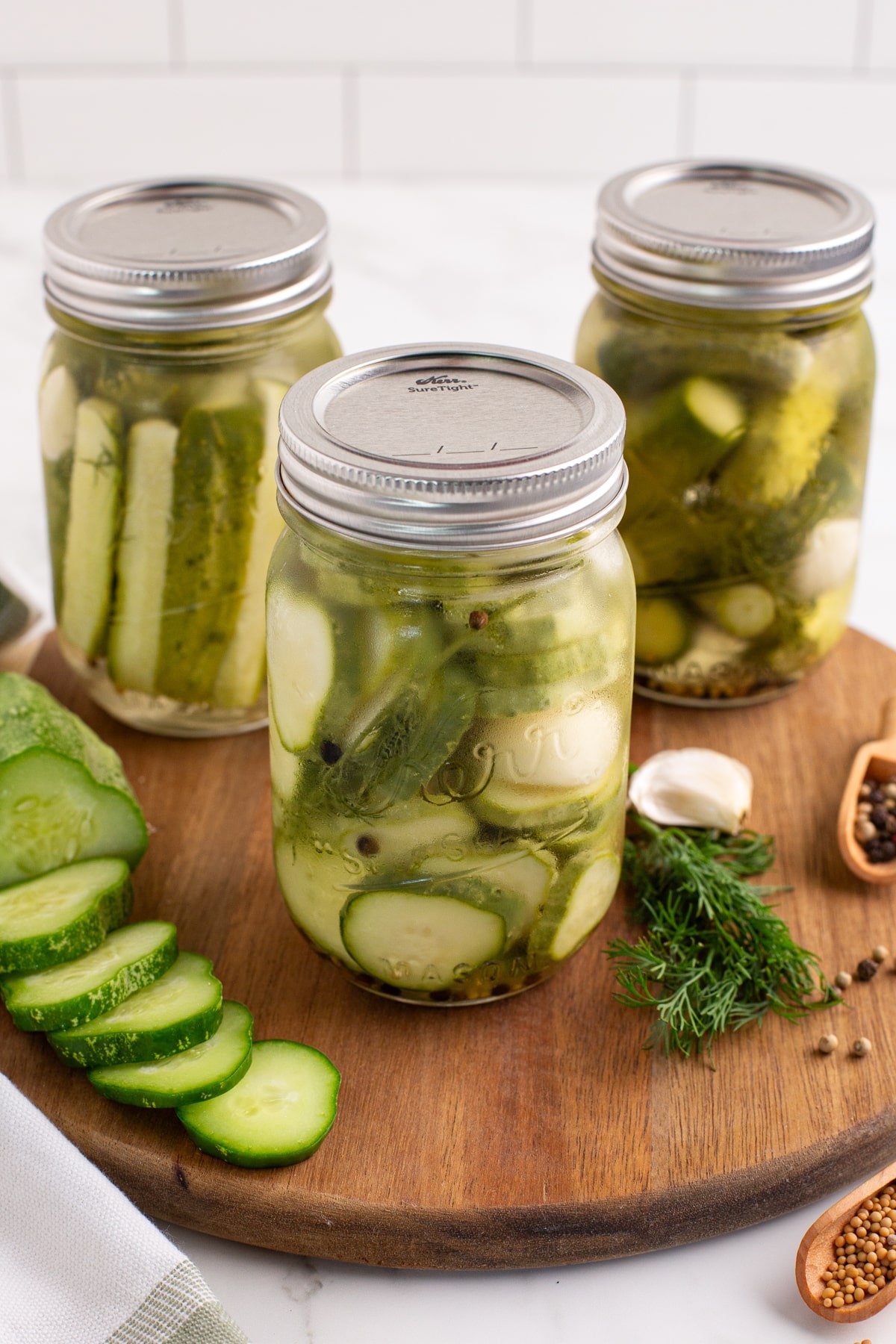 3 jars of pickled cucumbers on a wood cutting board.