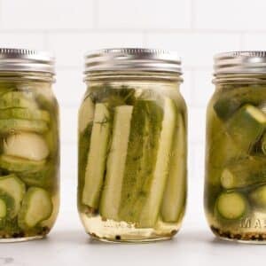 Three jars of pickles against a white tile background. Includes dill pickle chips, spears, and whole pickles.