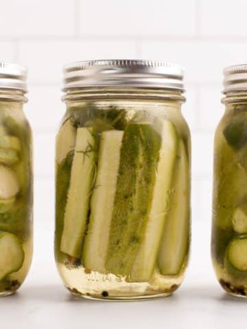 Three jars of pickles against a white tile background. Includes dill pickle chips, spears, and whole pickles.