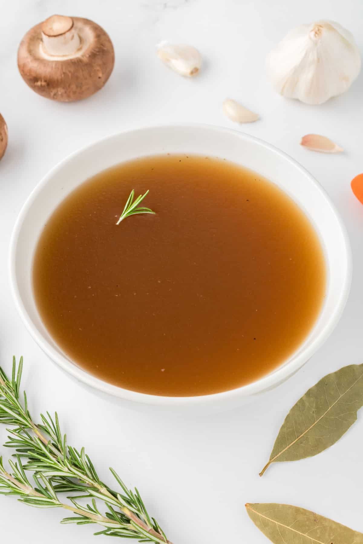 A bowl of clear beef broth, garnished with a tiny piece of fresh rosemary.
