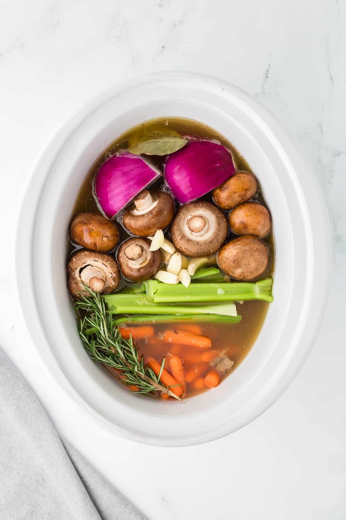 A white slow cooker insert filled with broth, fresh onions, mushrooms, celery, carrots, and fresh rosemary.