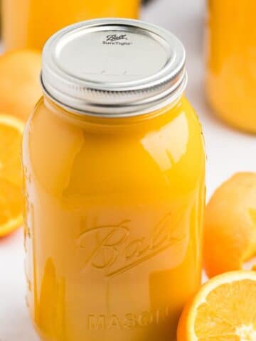 A mason jar filled with orange juice; fresh oranges surround the counter in the background.