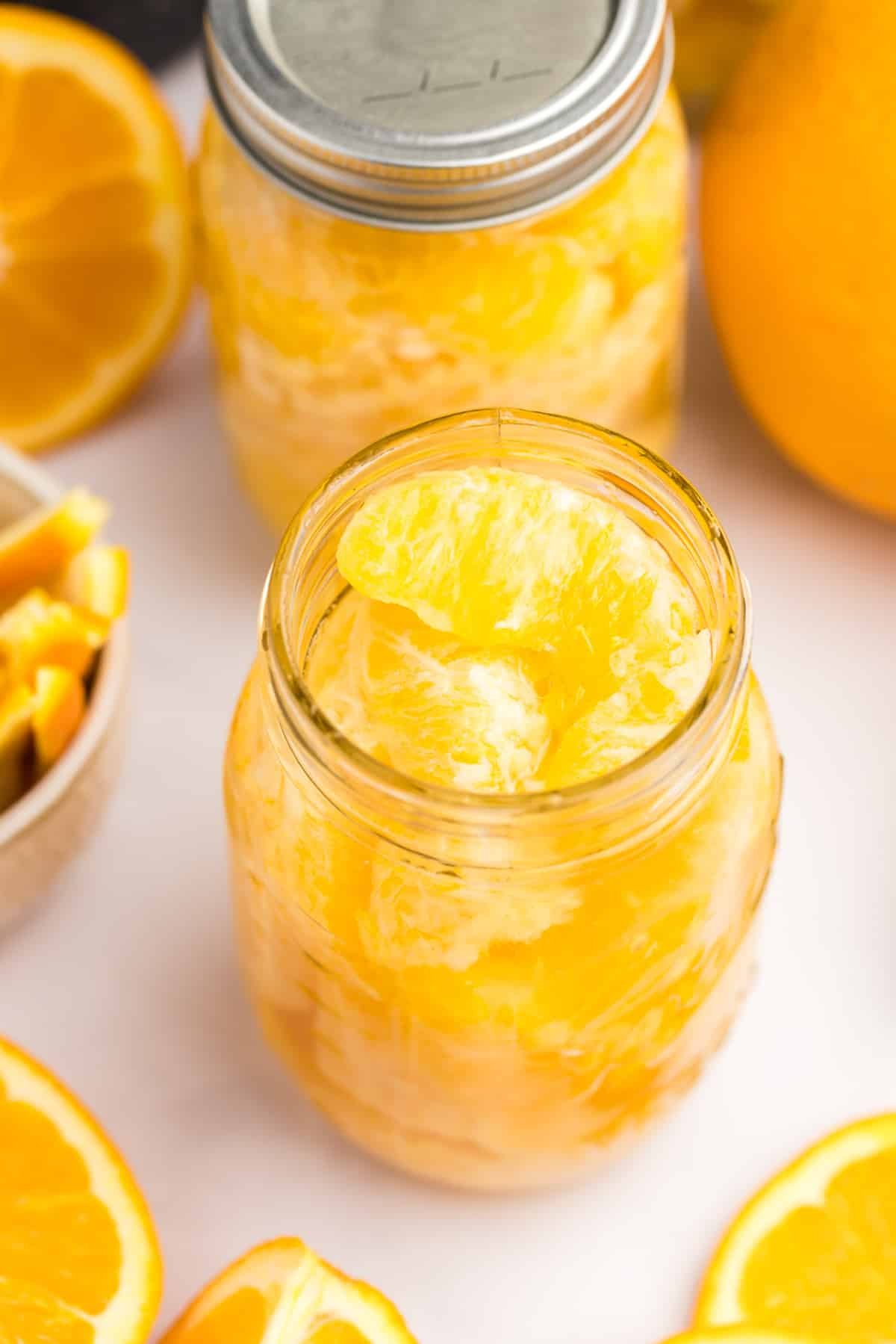 Jars of home canned oranges, surrounded by fresh oranges in the background.