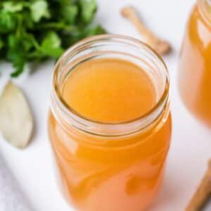 A glass mason jar filled with broth, with a bunch of parsley in the background.