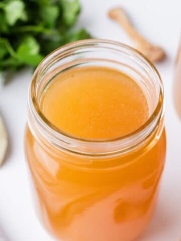 A glass mason jar filled with broth, with a bunch of parsley in the background.