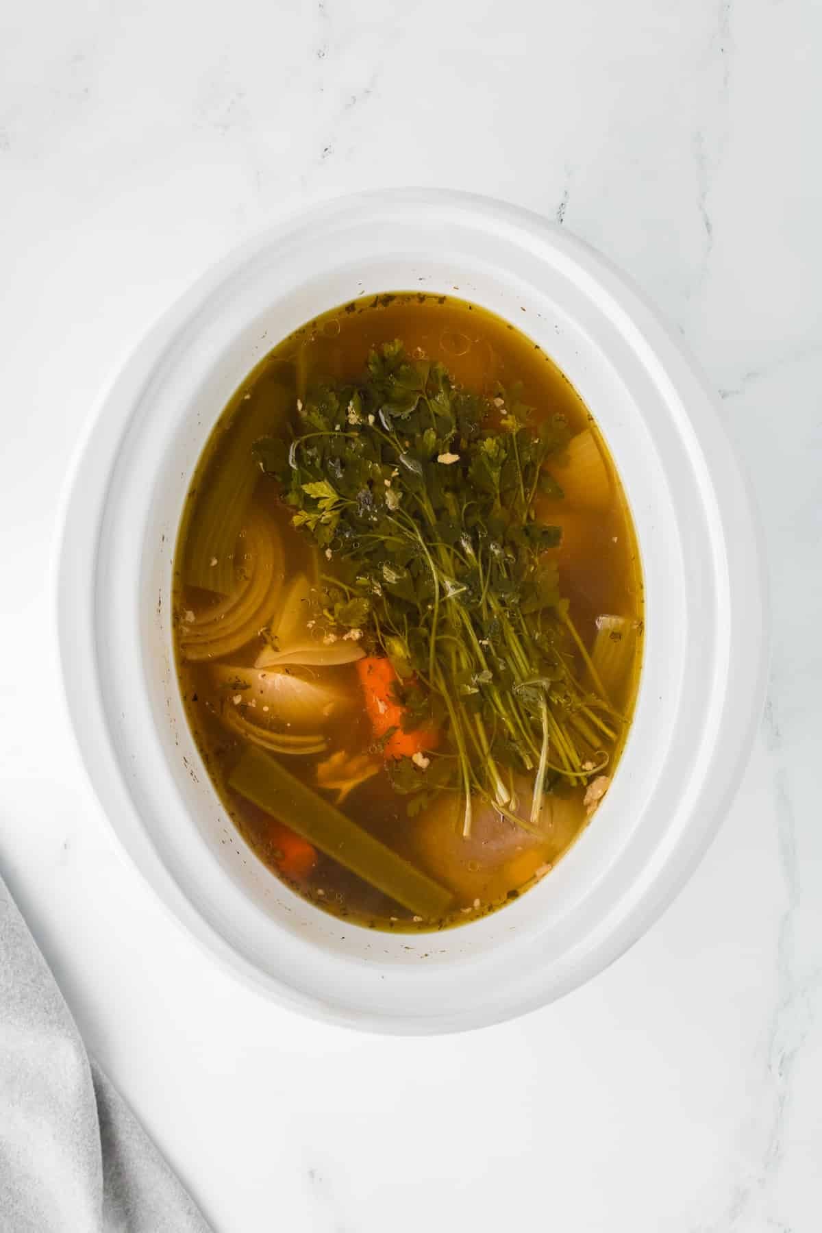 A slow cooker filled with broth with vegetables and parsley.