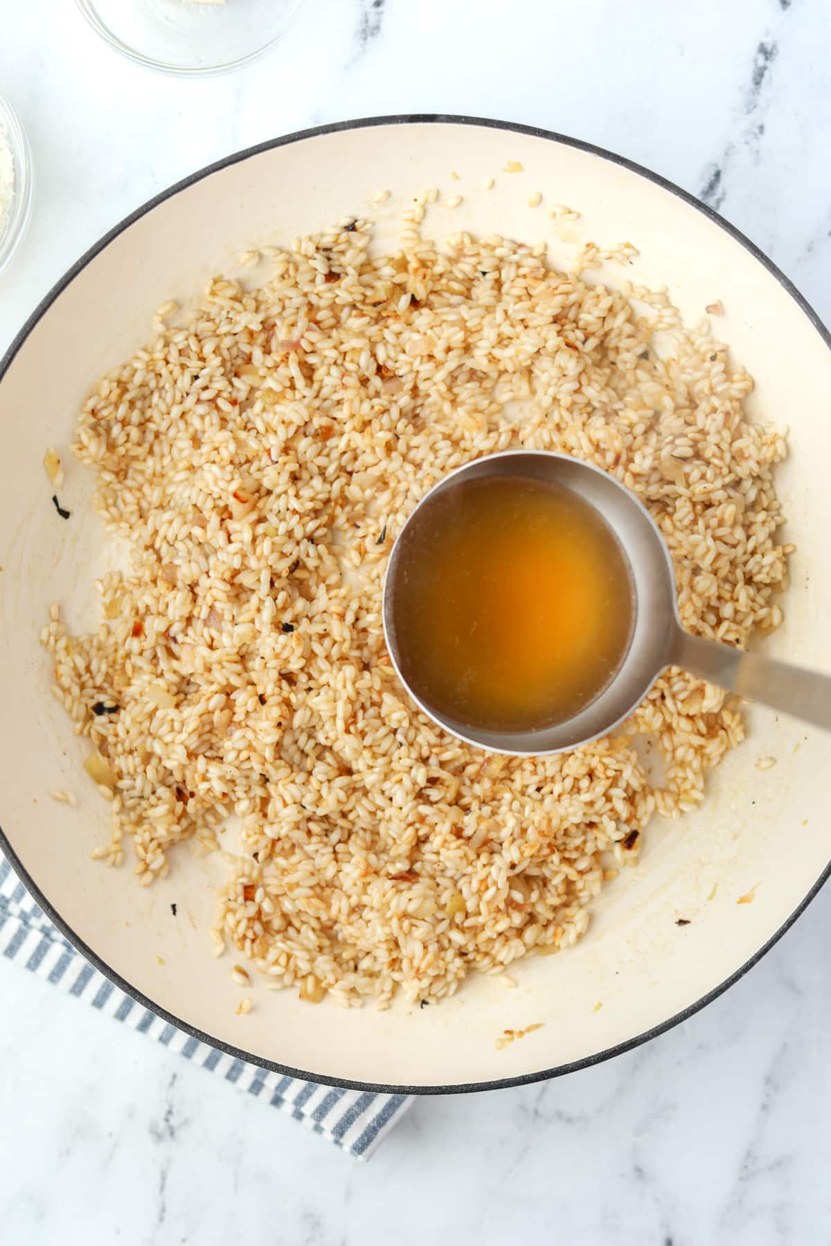 Ladling hot broth into an enameled skillet filled with toasted rice.