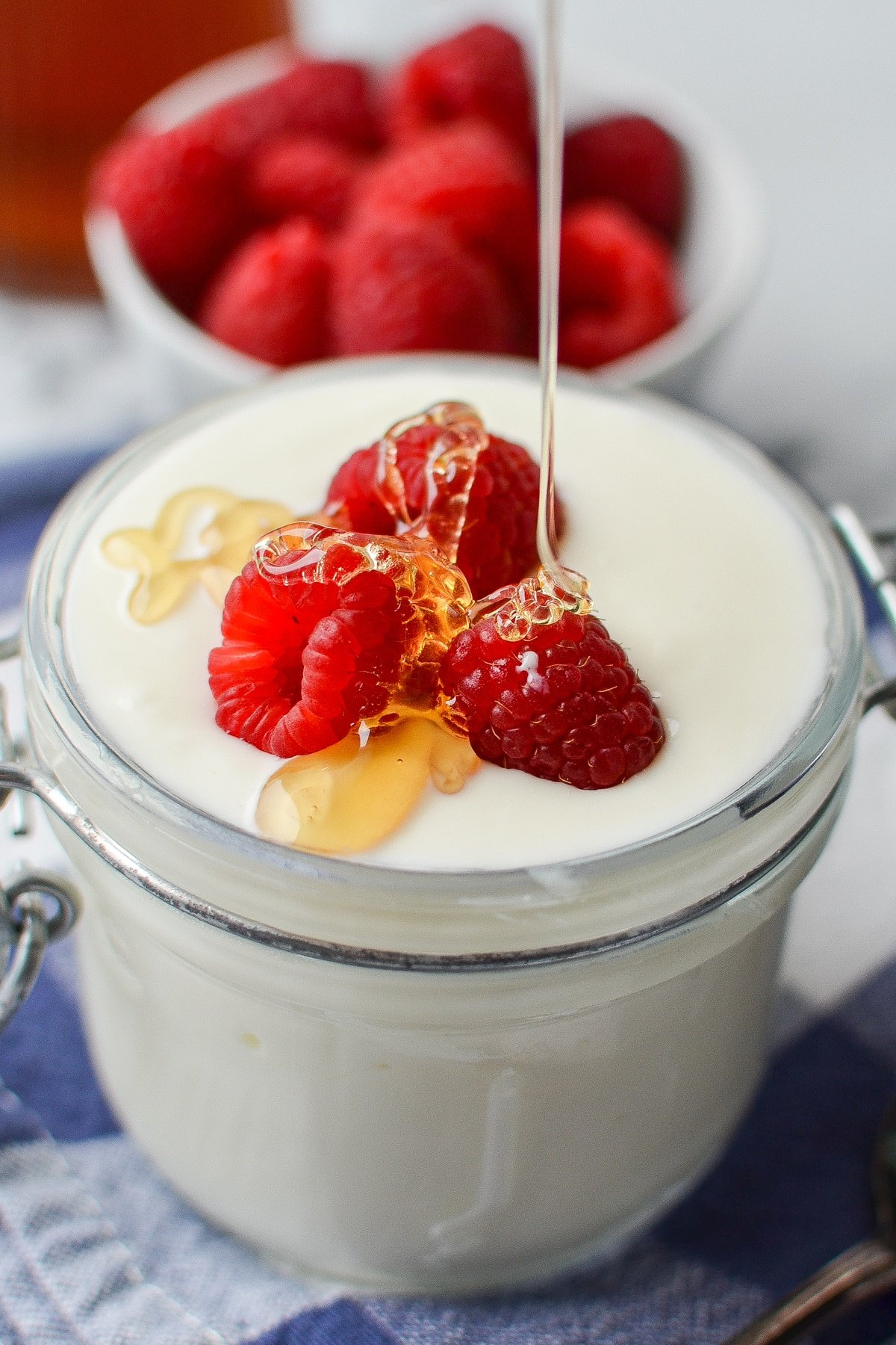 A glass jar filled with yogurt, topped with raspberries, and being drizzled with honey.