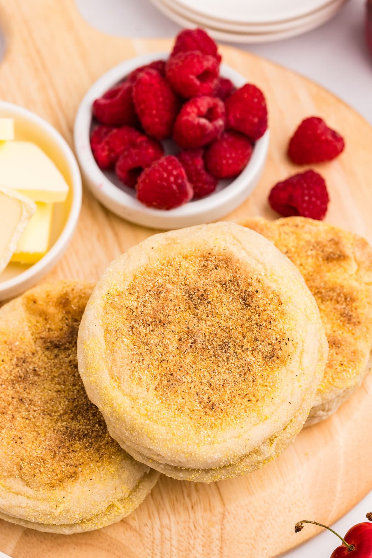 A stack of english muffins on a cutting board, with a small bowl of raspberries on the side.