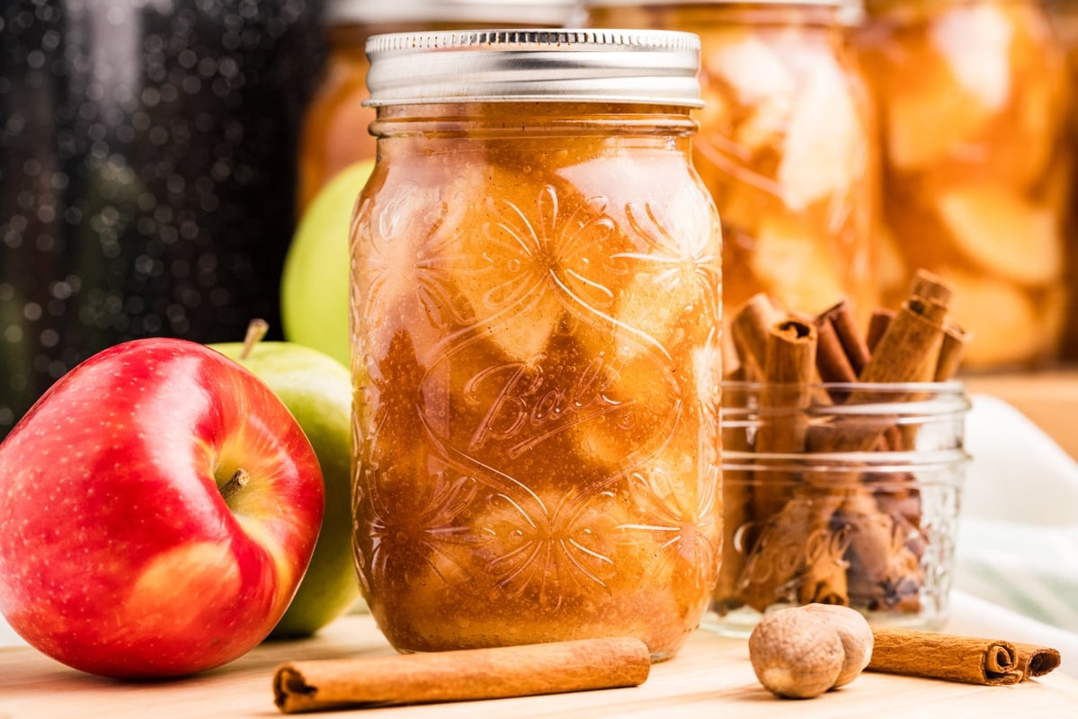 A jar of apple pie filling in front of a water bath canner, surrounded by apples and cinnamon sticks.