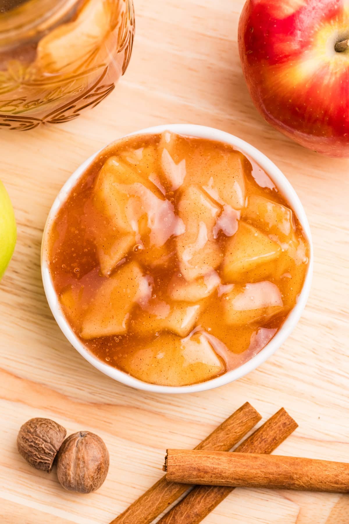 A small bowl filled with a sweet apple pie filling, surrounded by spices, apples, etc.