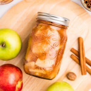 A canning jar of apple pie filling laying on its side, with apples and cinnamon around the perimeter.