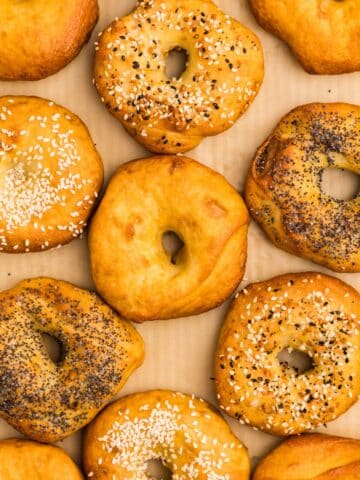 Many bagels arranged on a baking sheet, covered with various toppings.