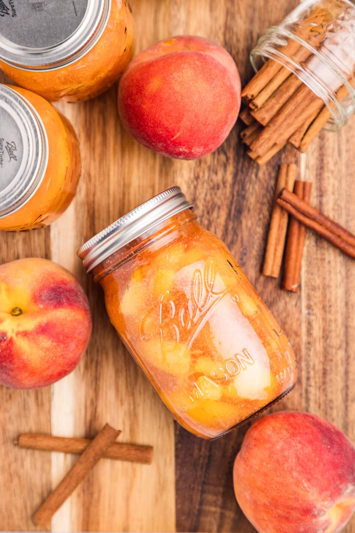 A jar of peach pie filling surrounded by cinnamon sticks and fresh peaches.