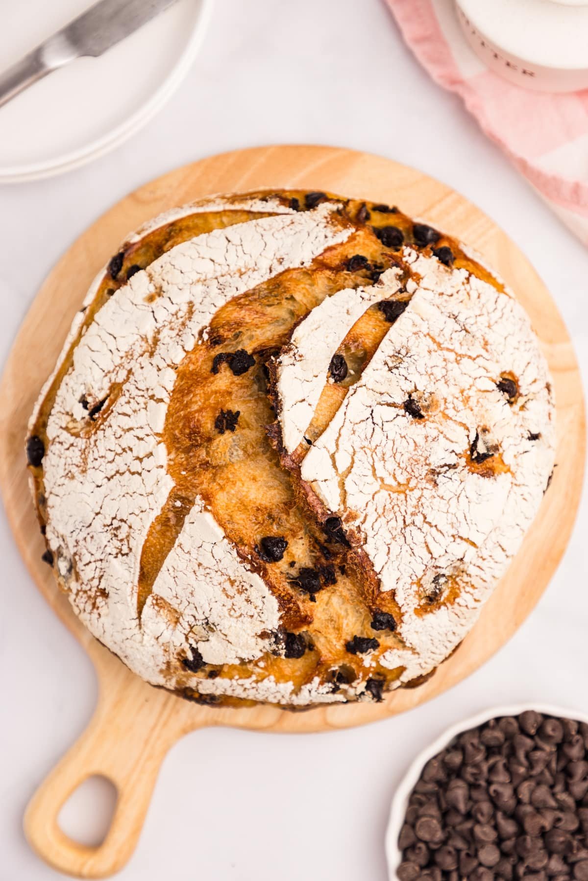 A loaf of sourdough bread on a round cutting board and dotted with chocolate chips.