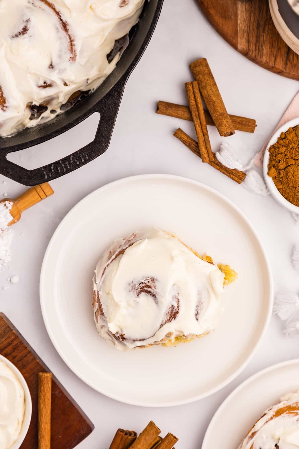 A cinnamon roll on a white plate, surrounded by cinnamon sticks, and a full pan of cinnamon rolls.